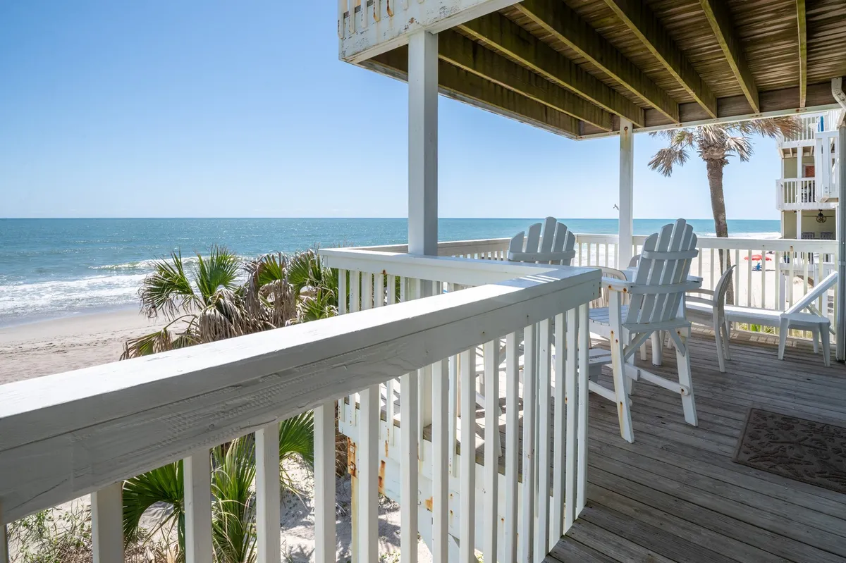 The image showcases an oceanfront condo deck at Kure Beach, with classic Adirondack chairs perfectly positioned to take in sweeping Atlantic views. Palm trees frame the scene, leading the eye to the sparkling shoreline. The tranquil setting invites relaxation, whether enjoying a morning coffee at sunrise, unwinding with a good book, or sharing a sunset cocktail with friends.