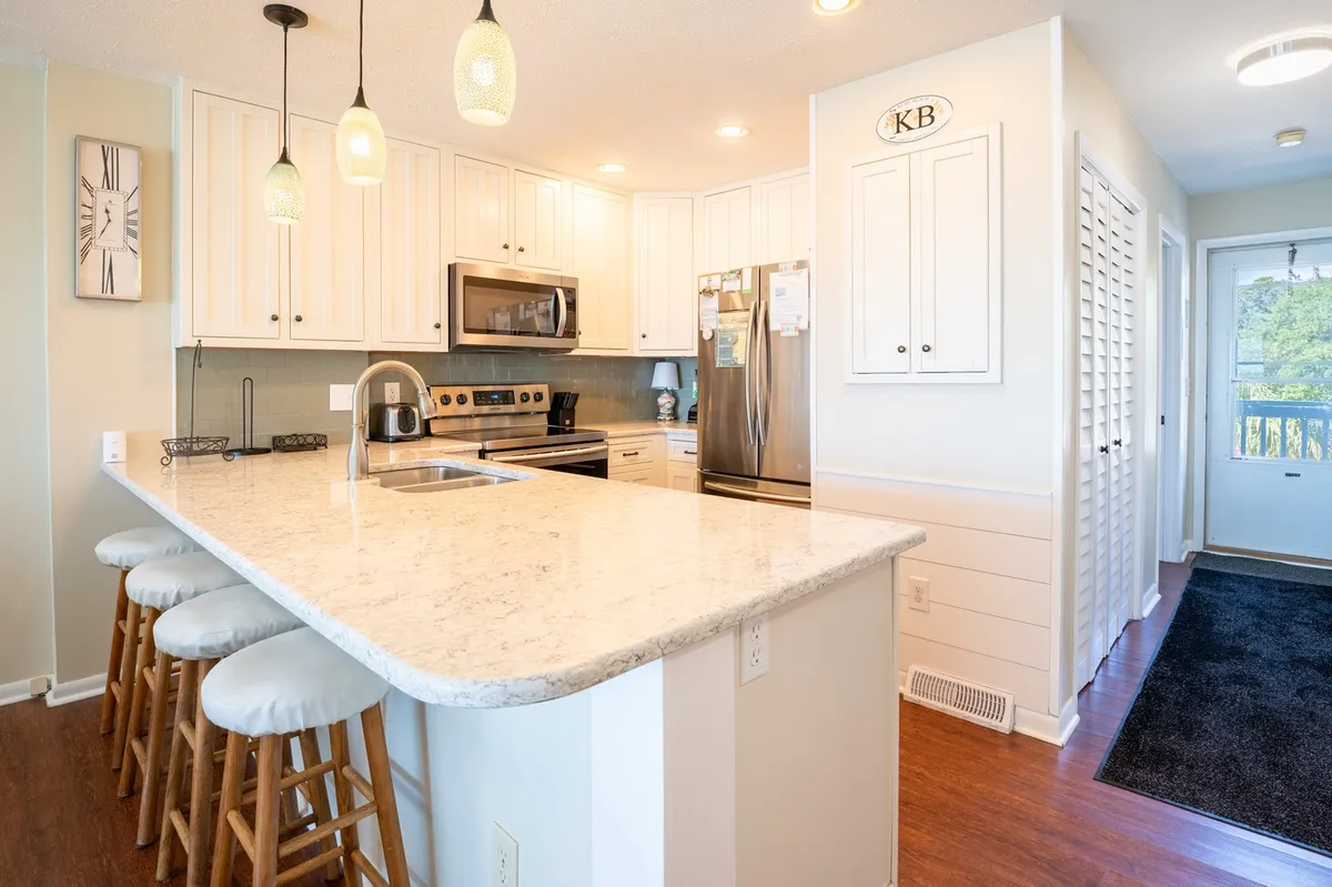 The image features a bright kitchen with a central island and bar seating for four. Stools line the island, making it an inviting spot for casual dining or conversation. Large windows and modern appliances enhance the inviting mood, while the kitchen opens out to the adjacent living and dining areas.