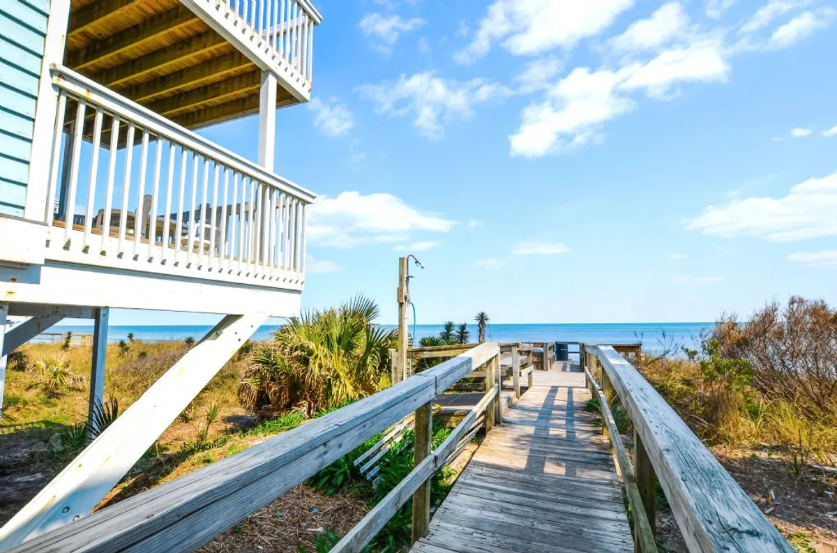 A tranquil wooden boardwalk leads guests across beach vegetation towards a wide ocean view, with the house balcony visible. The scene captures the proximity to the beach and the beautiful natural surroundings, inviting exploration and seaside adventure.