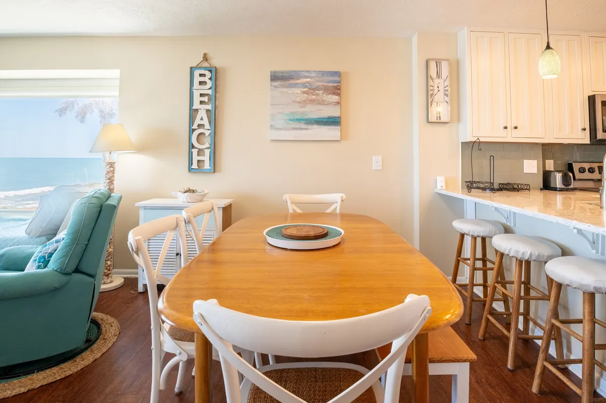 This image showcases a dining table set for six with a direct view of the beach through expansive windows. The dining area is open to the kitchen, and decorative art on the walls adds warmth. The space is filled with natural light, highlighting the casual and inviting setting ideal for family meals and social gatherings.