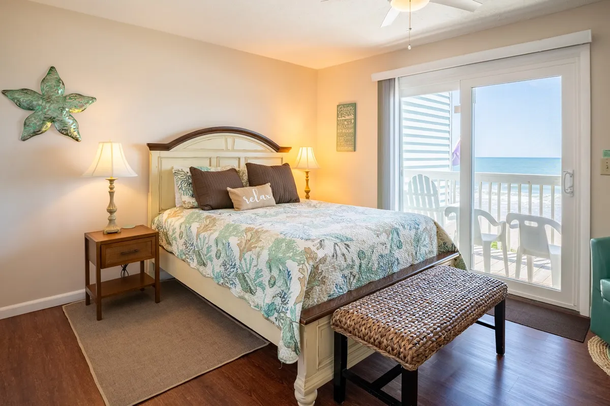 This image features a bed with floral bedding situated near a sliding door that opens to an ocean view. Decorative accents like a starfish and lamps add a soothing, coastal touch. Natural light streams in, highlighting the tranquil and refreshing mood of the master suite.