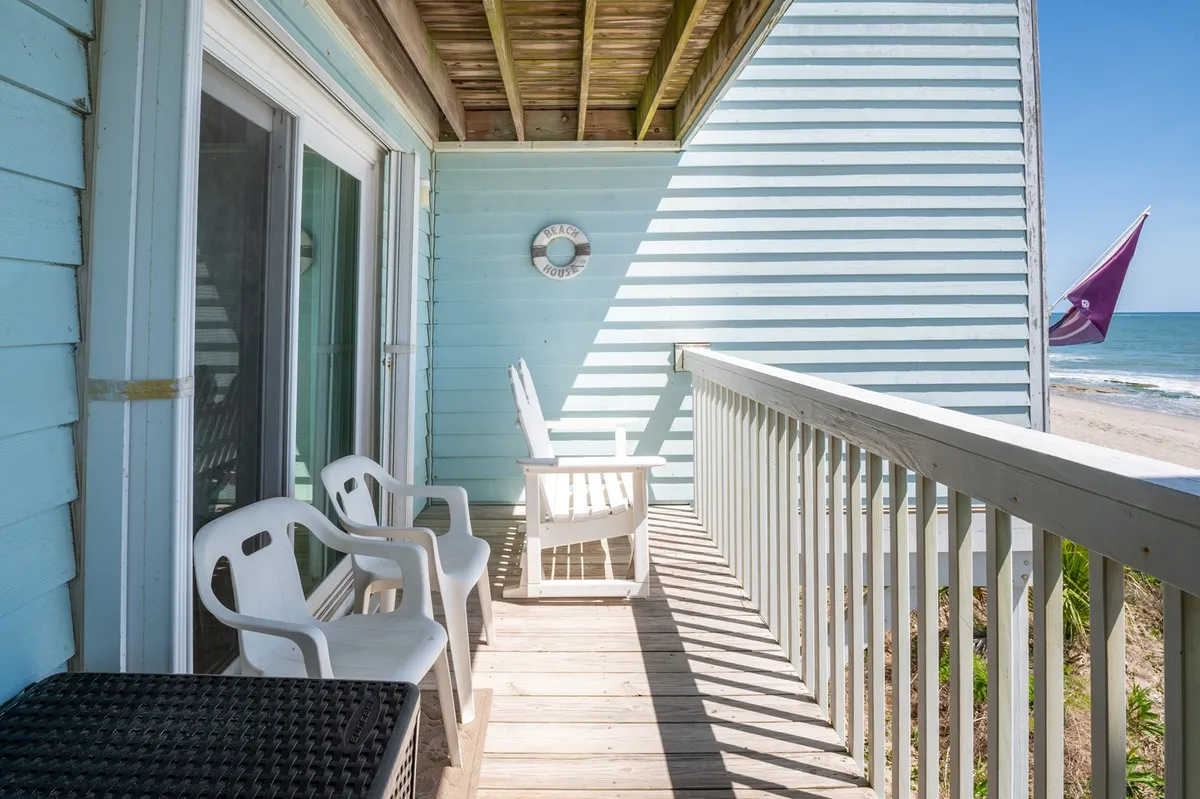 A serene outdoor setting with white chairs facing the beach, the gentle arc of the shoreline visible beyond the sand dunes. The beach house exterior and decorative wall clock add character, while soft lighting suggests either dawn or dusk, emphasizing peacefulness and reflection.