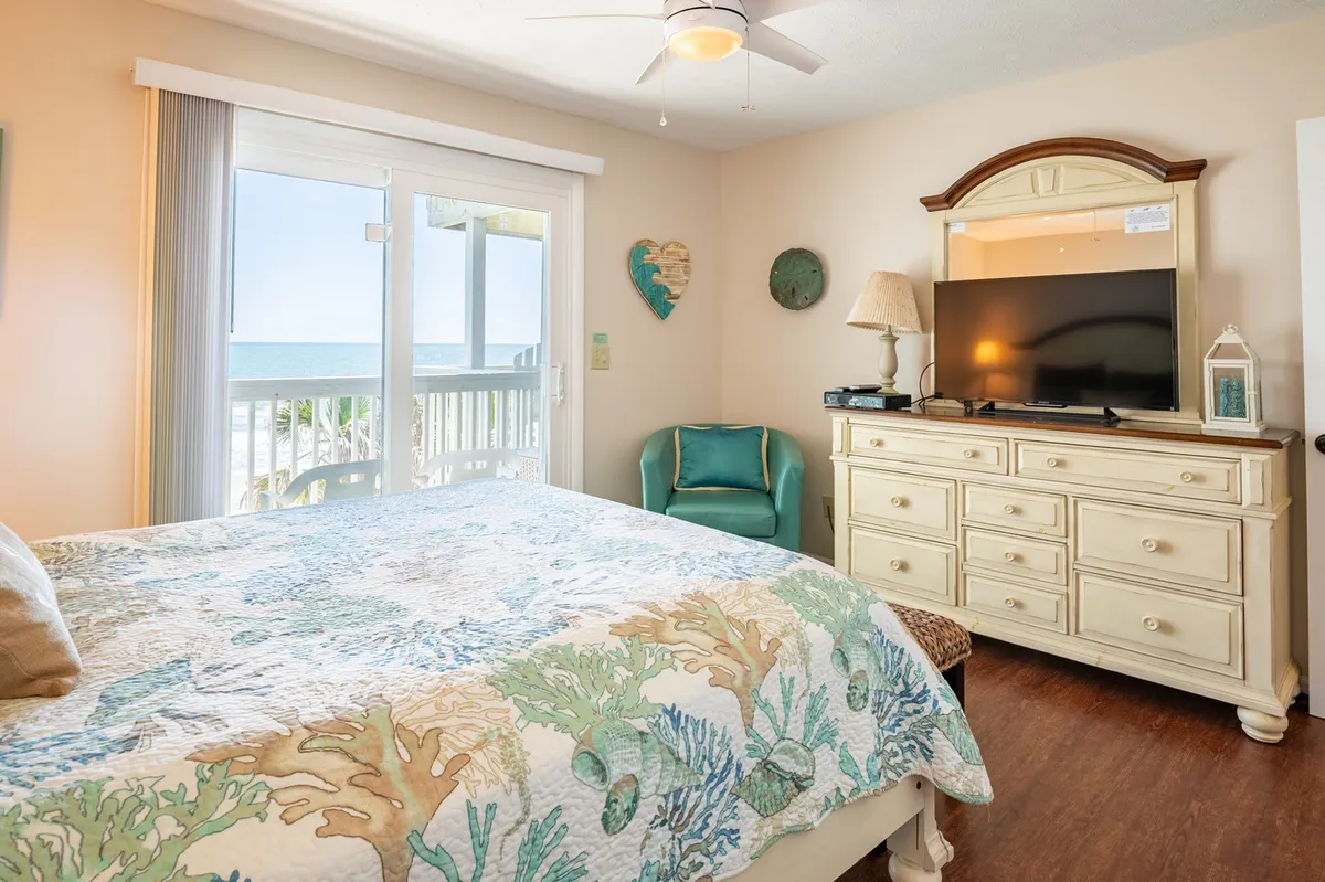 A cozy bedroom with a neatly made bed featuring a colorful quilt, sliding glass doors leading to a balcony, and a TV cabinet. The room is filled with natural light, presenting a fresh and inviting atmosphere.