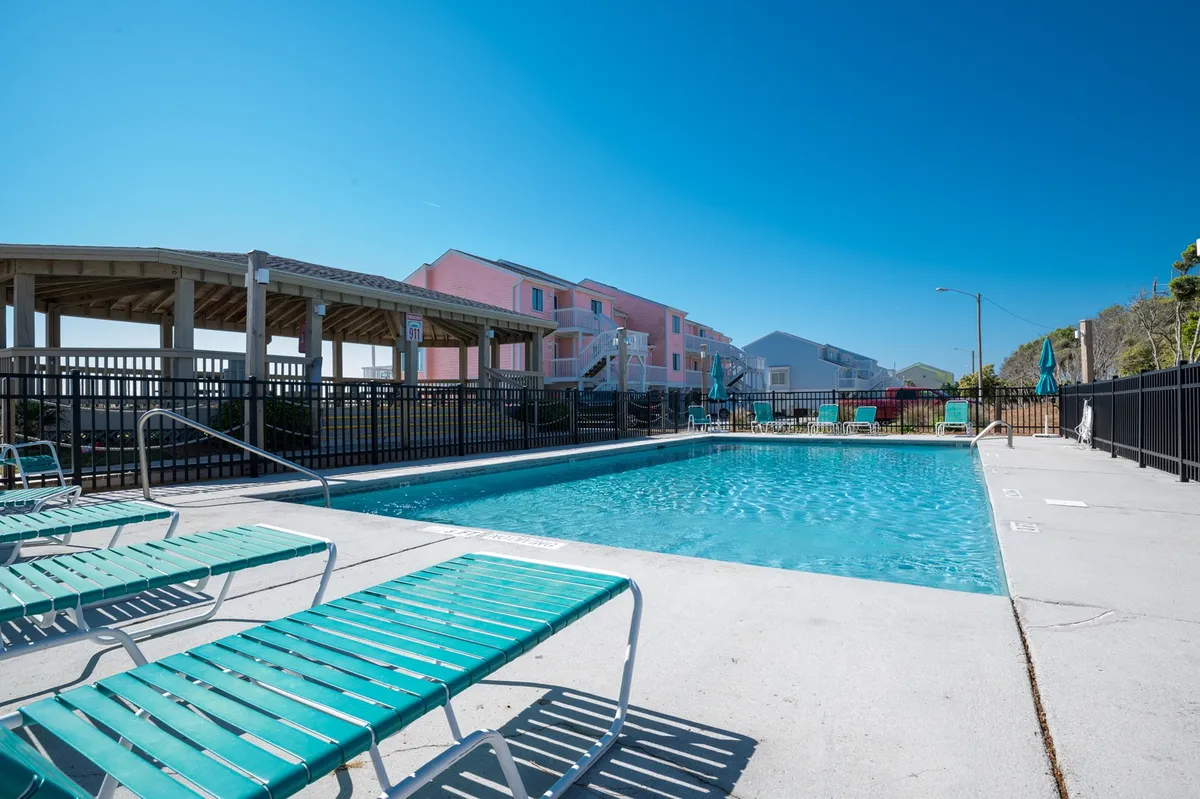 A sparkling outdoor swimming pool surrounded by lounge chairs and bordered by resort buildings. The scene captures a sunny, relaxed atmosphere, ideal for socializing, swimming, or unwinding after a day at the beach.