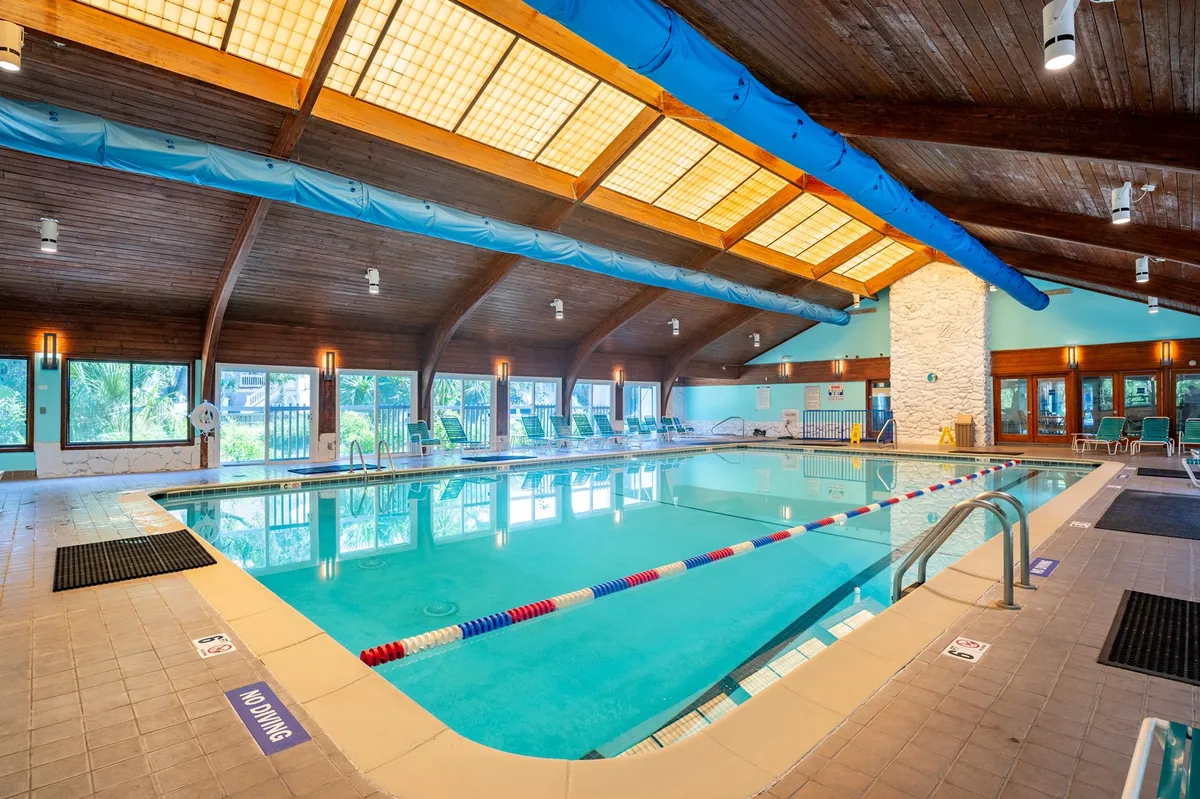 An inviting indoor swimming pool illuminated by natural light streaming through large windows. Wooden ceilings add warmth, while lounge chairs line the pool, providing comfortable spots for relaxation. The environment is bright, tranquil, and perfect for year-round recreation.