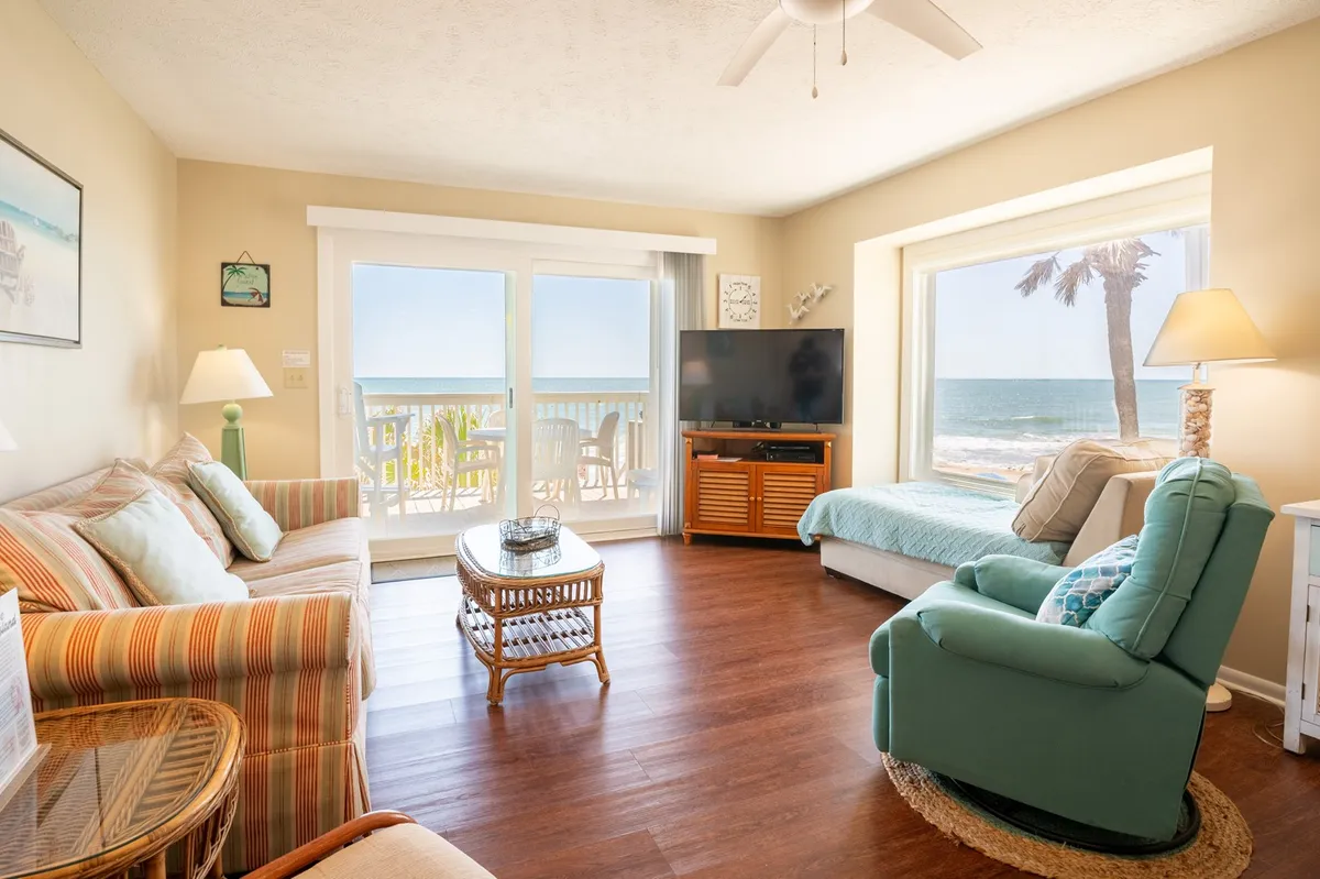 This image features a spacious living area with large windows framing a stunning ocean view. The room includes a comfortable seating area with plush sofas and a television, as well as tasteful decorative touches on the walls. Natural light fills the space, and the layout suggests a welcoming environment for relaxation and socializing, with direct visual access to the outdoors.