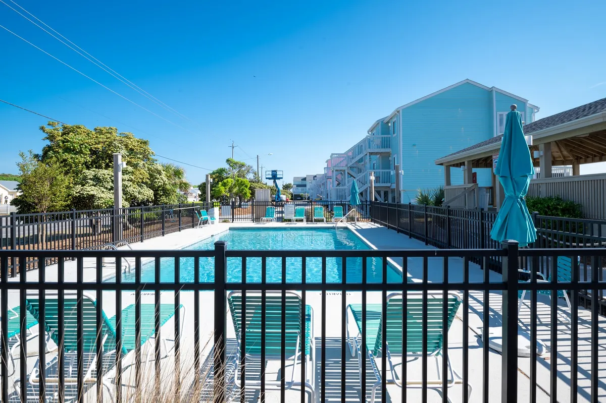 A sparkling swimming pool flanked by multiple lounge chairs and umbrellas, all enclosed within a safe fence. The open sky and relaxed setting invite groups to swim, sunbathe, or socialize poolside, showcasing the resort-style amenities available to guests.
