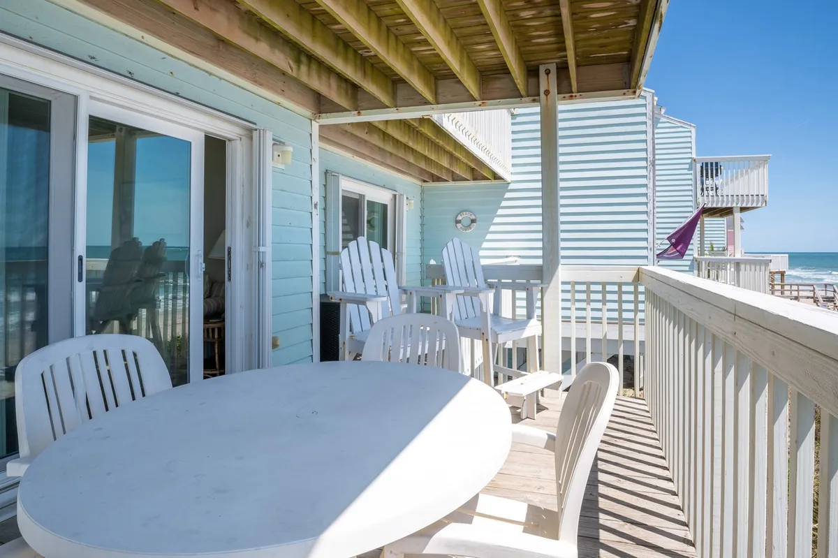 An oceanfront deck features a spacious outdoor dining table surrounded by white chairs, set against a sweeping view of the beach and blue water. The scene is tranquil, inviting guests to gather for meals or sunset conversations while enjoying the sound and breeze of the ocean.