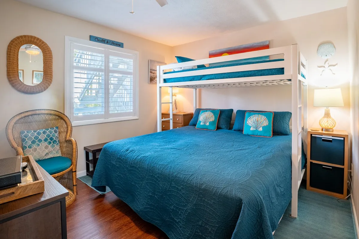 A cozy bedroom featuring a large bed paired with a twin loft bunk, a woven chair, and a side table with a lamp. Sunlight filters through the shutters, highlighting a relaxed and inviting environment ideal for families or friends.