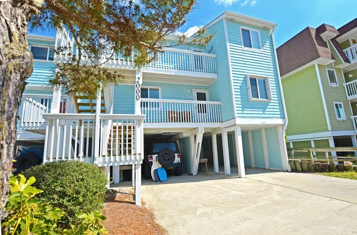The image displays the blue exterior of the house with visible balconies and landscaping. There is a driveway area where vehicles can be parked, and the general scene conveys a welcoming approach to the property.