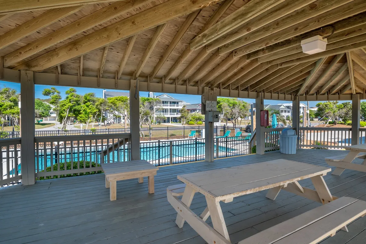 An exterior shot of a communal area with wooden picnic tables, open beams, views of the pool, and surrounding greenery. The setting is casual and sociable, perfect for family barbecues, gatherings, or a quick break between activities.