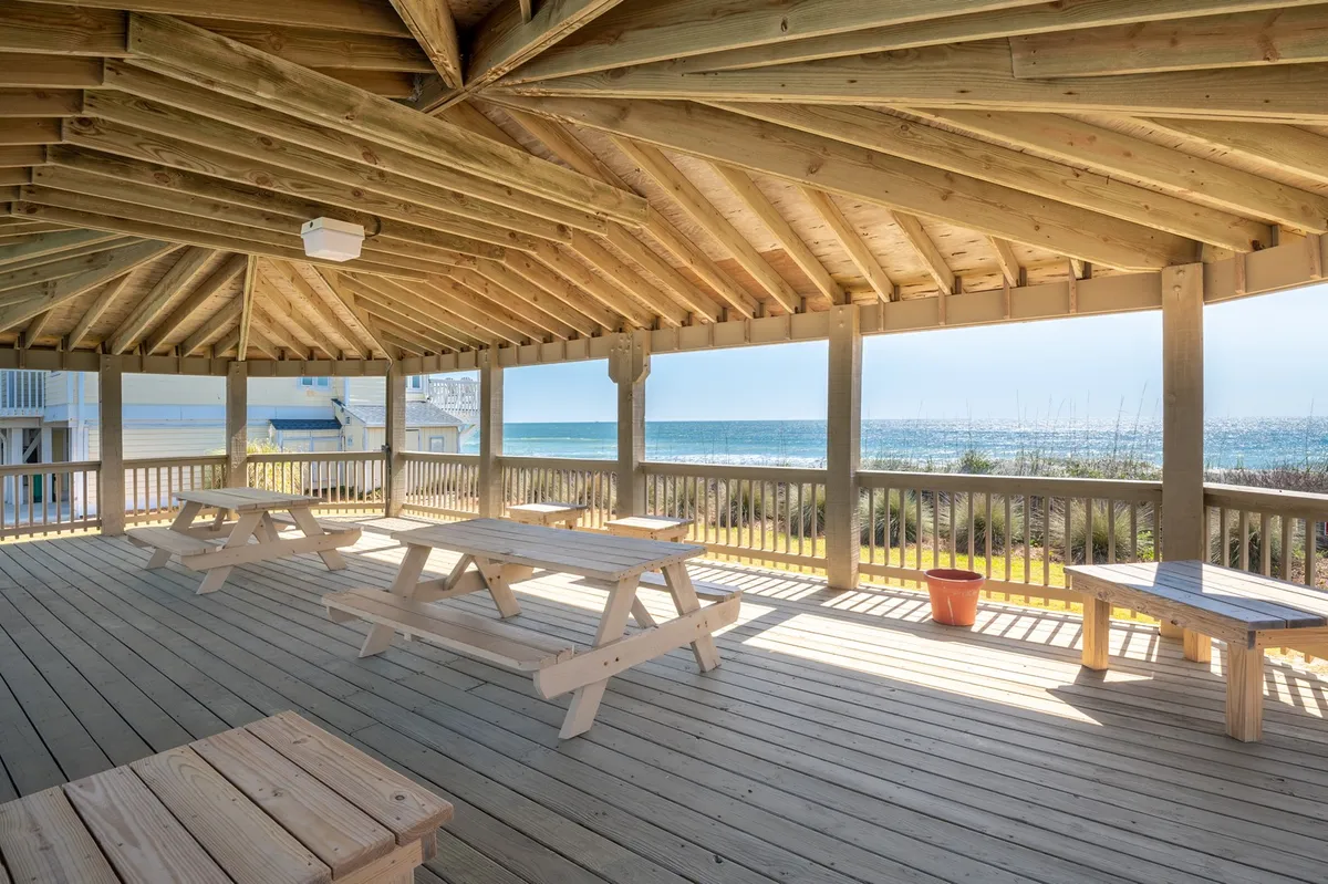 A covered outdoor picnic area with rustic wooden tables overlooks the ocean, creating a perfect spot for casual dining, gathering with friends, or simply watching the sunset. The open-air design emphasizes the natural coastal setting and accessibility to outdoor experiences.